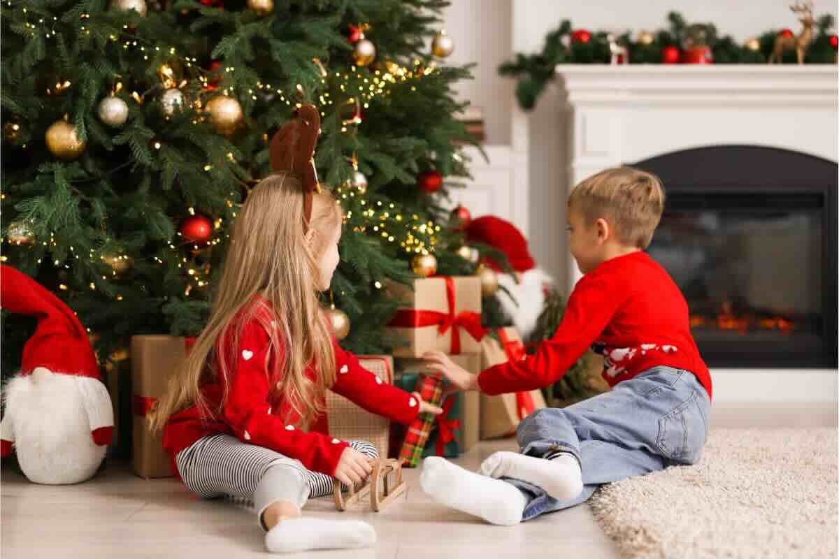 Due bambini giocano con i regali sotto l'albero di Natale, vestiti con maglioni rossi natalizi