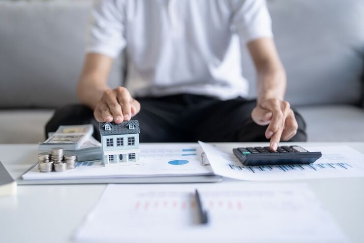 Uomo fa calcolo alla calcolatrice. Sulla scrivania documenti e una riproduzione di una casa, e una penna 